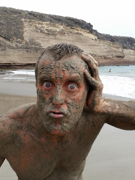 Foto ritratto di un uomo coperto di fango sulla spiaggia contro il cielo