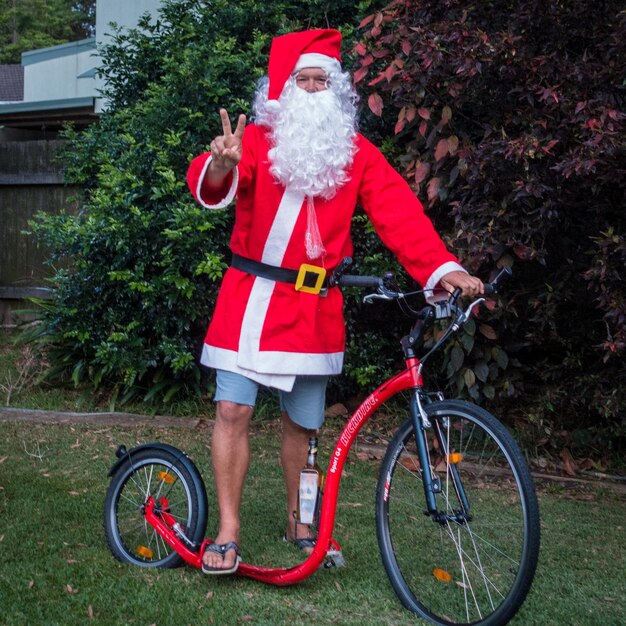 Foto ritratto di un uomo vestito da babbo natale che mostra un segno di pace su una bicicletta nel cortile sul retro