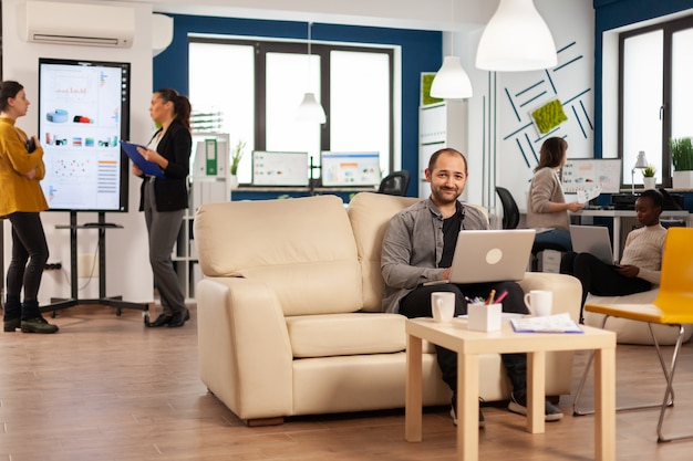 Portrait of man employee typing on laptop looking at camera smiling while diverse team working in background