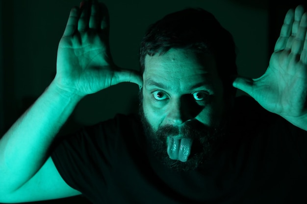 Photo portrait of man gesturing while sticking out tongue against black background