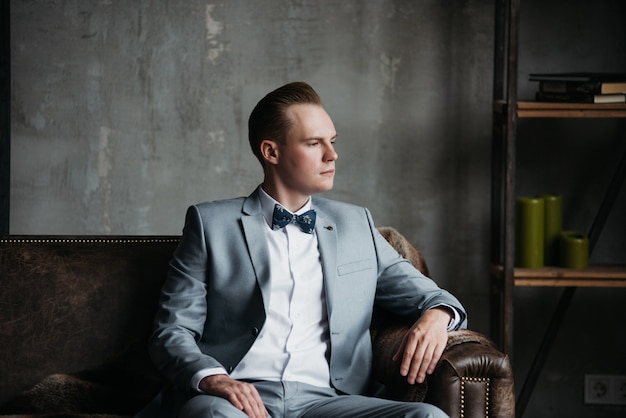 Portrait of a man. The groom in a in a gray suit, white shirt and a bow tie is sitting on a brown leather couch. Room in loft style.