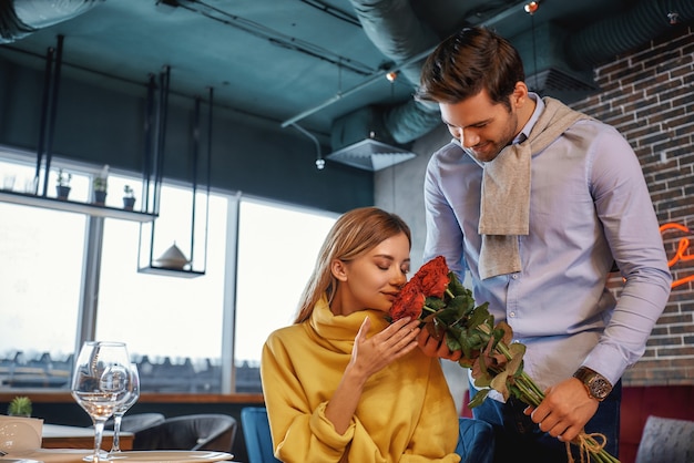 Portrait of man holding a bouquet of roses while standing in the restaurant. Woman smells the flowers. Romantic dinner concept. Horizontal shot