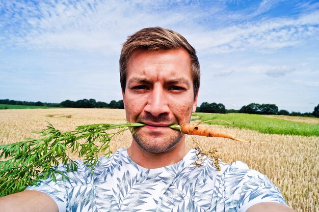 Photo portrait of man holding carrot in mouth on agricultural field against sky