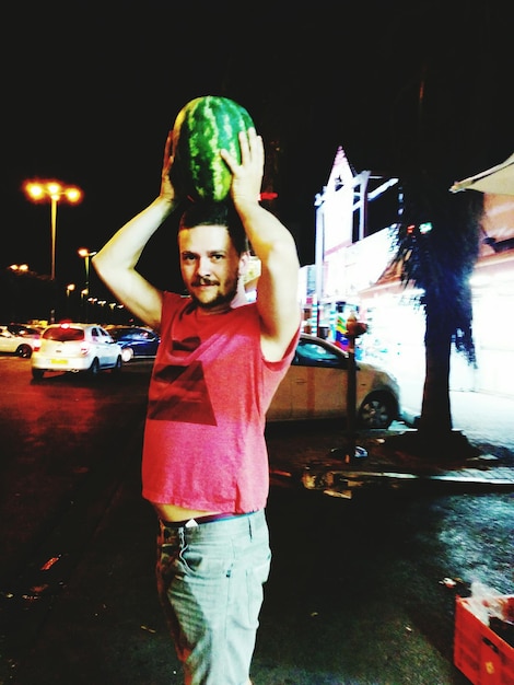 Photo portrait of man holding watermelon on head by street at night