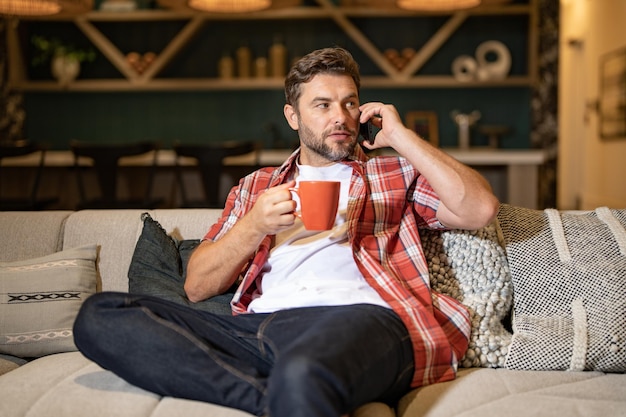 Portrait of man looking at smart phone at living room man is talking hold cup of coffe chatting on