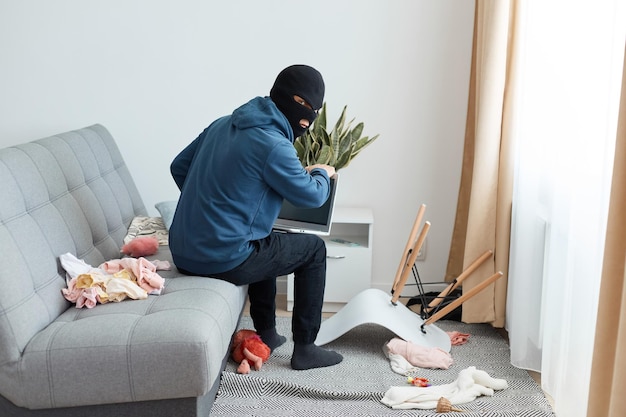 Portrait of man robber wearing blue hoodie and black balaclava sitting on the couch backwards to the camera, turning around to see that no one is coming, stealing a laptop.