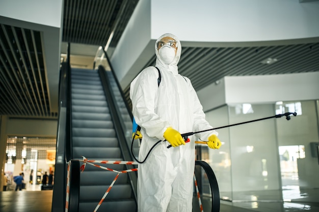 Portrait of a man in a sanitizing disifection suit holding\
spray near the escalator in an empty shopping mall. a volunteer\
cleaning up the public places to prevent covid-19. health awareness\
concept.