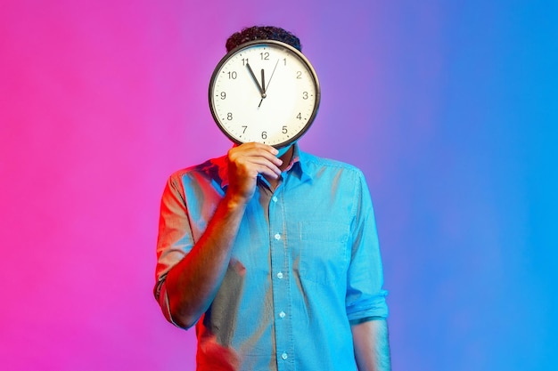 Portrait of man in shirt hiding face behind big wall clock display, wasting his time, procrastination, bad organization of working time. Indoor studio shot isolated on colorful neon light background.