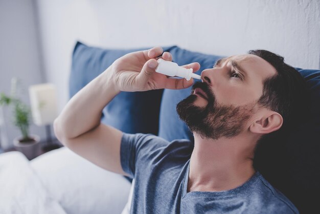 Portrait of man sitting in the bed spraying drops in his nose