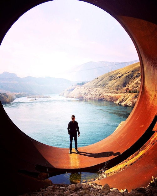 Foto ritratto di un uomo in piedi su un grande tubo a cerchio con il fiume sullo sfondo alla diga
