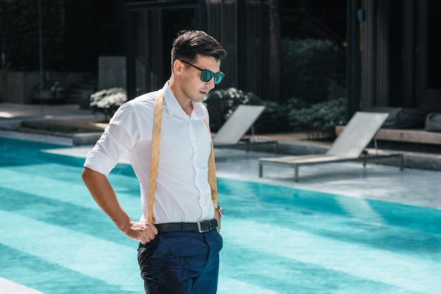 Portrait of the man in sunglasses standing in the swimming pool wearing a wet white shirt