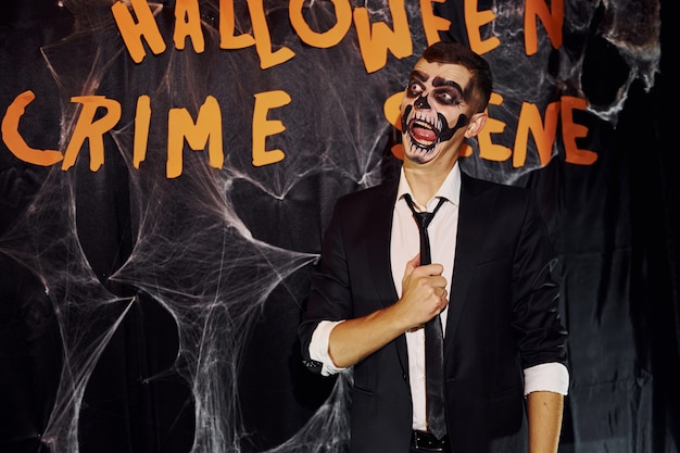 Photo portrait of man that is on the thematic halloween party in scary skeleton makeup and costume against crime scene wall.