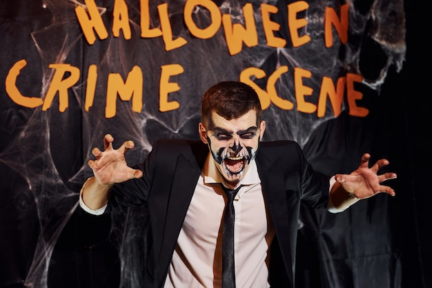 Portrait of man that is on the thematic halloween party in scary skeleton makeup and costume against crime scene wall.