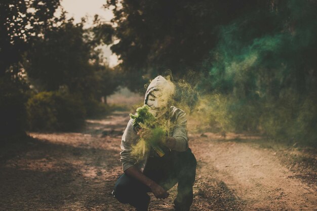 Foto ritratto di un uomo che indossa una maschera mentre tiene in mano una torcia di soccorso su una strada di terra tra gli alberi