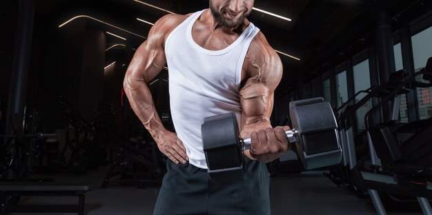 Portrait of a man in a white T-shirt exercising in the gym with dumbbells. Biceps pumping. Fitness and bodybuilding concept.