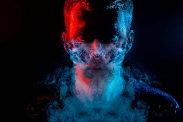 Portrait of a man who is angry and has bad emotions with mask from a smoke around on a black isolated background The face of the guy in the shade with color red and blue illumination