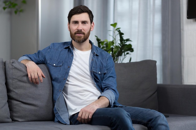 Portrait of a man with a beard at home sitting on the couch and looking at the camera