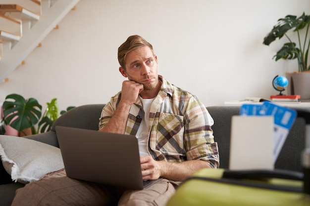 Photo portrait of man with concerned face looks aside with worried expression sits on sofa with laptop has