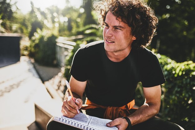 Portrait of man with curly hair writing some notices on paper and preparing for exams wearing black tshirt sitting on the city street Freelancer businessman makes plans for new projects