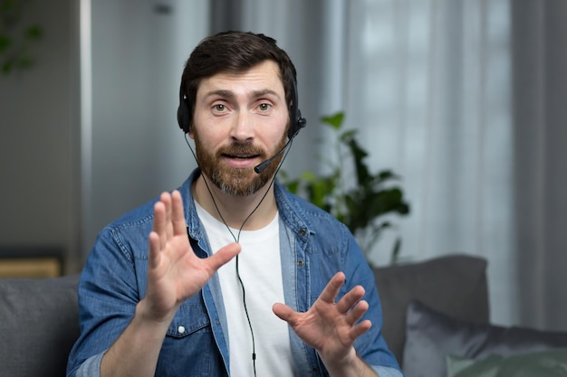 Portrait of a man with a headset working online consultant at home focused and angry talking to a client looking at the webcam