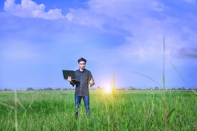 Portrait of man with laptop gesturing while standing on field against blue sky