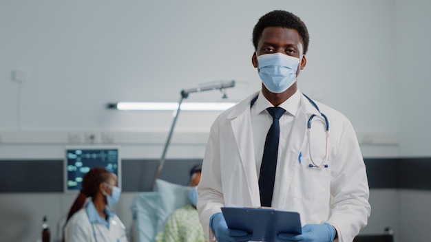 Portrait of man working as doctor with stethoscope and files, standing with face mask in hospital ward. General practitioner with white coat and checkup documents during pandemic.