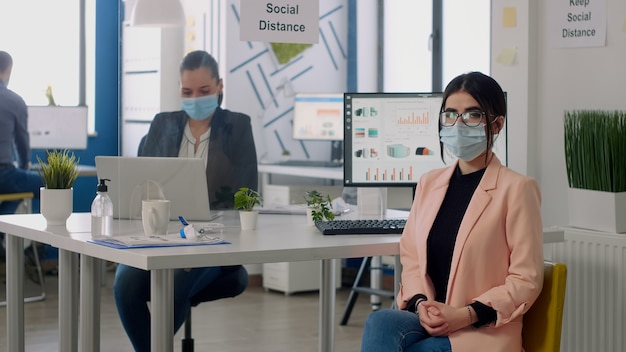 Portrait of manager with face mask sitting on chair at desk table in nre normal business company office. Teamworkers working in background respecting social distancing during coronavirus pandemic