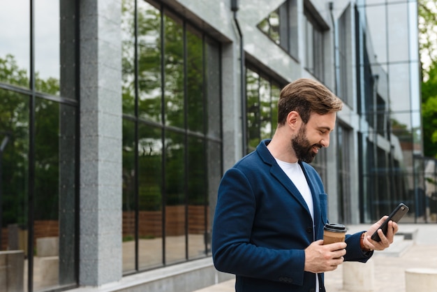 Portrait of masculine businessman in jacket holding mobile phone while standing outdoors near building with takeaway coffee