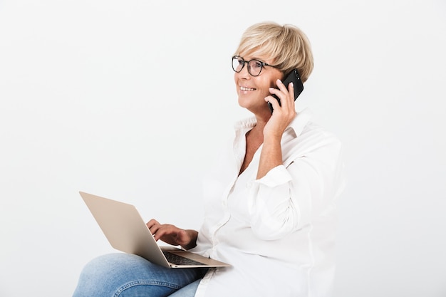 Portrait of mature adult woman wearing eyeglasses talking on smartphone while sitting with laptop computer isolated over white wall
