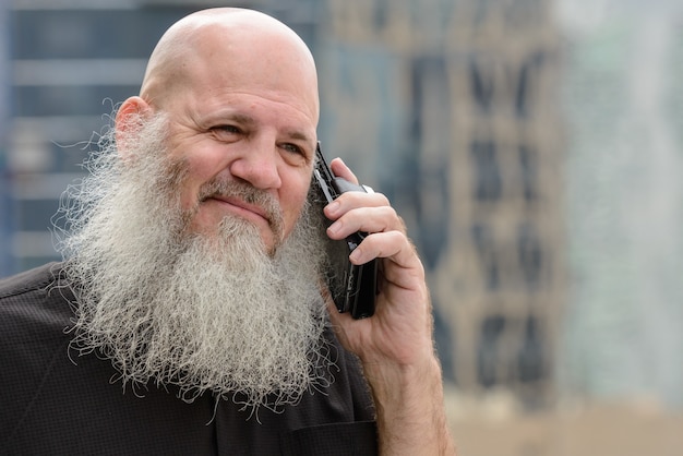 Portrait of mature bald hipster man with long beard against view of the city outdoors