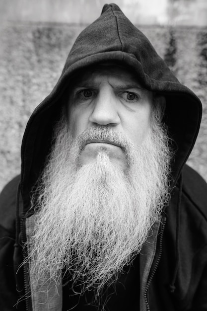 Portrait of mature bald man with long gray beard against grunge concrete wall outdoors in black and white