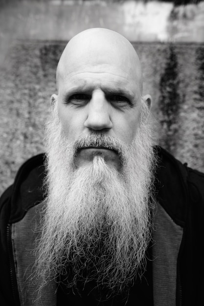 Portrait of mature bald man with long gray beard against grunge concrete wall outdoors in black and white