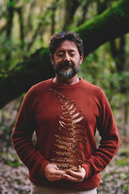 Photo portrait of mature bearded man closing eyes and holding dry leaf in the woods. concept of nature and environment. people loving woods and ambient. inner balance life with adult male