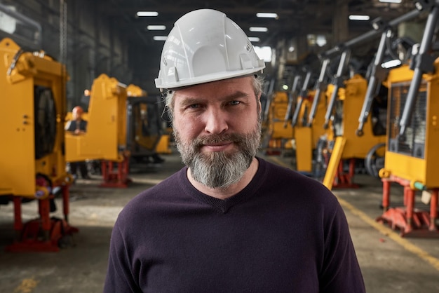 Portrait of mature bearded worker in work helmet looking at camera while standing in warehouse