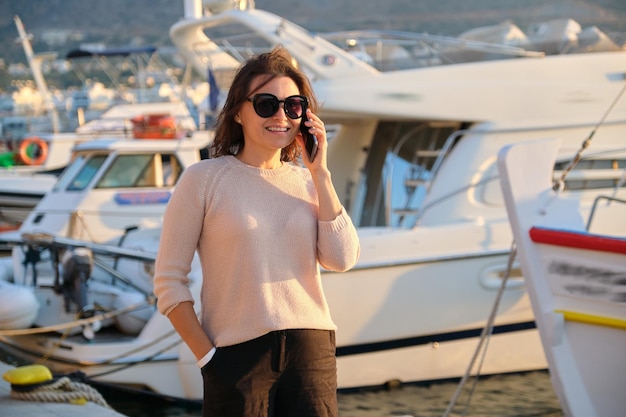 Portrait of mature beautiful woman walking in sea pier talking\
on mobile phone smartphone landscape mountains sunset at sea yacht\
club background