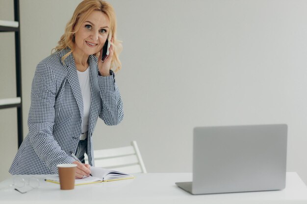 Portrait of a mature business woman sitting in a modern office talking on the phone and writing something in a notebook