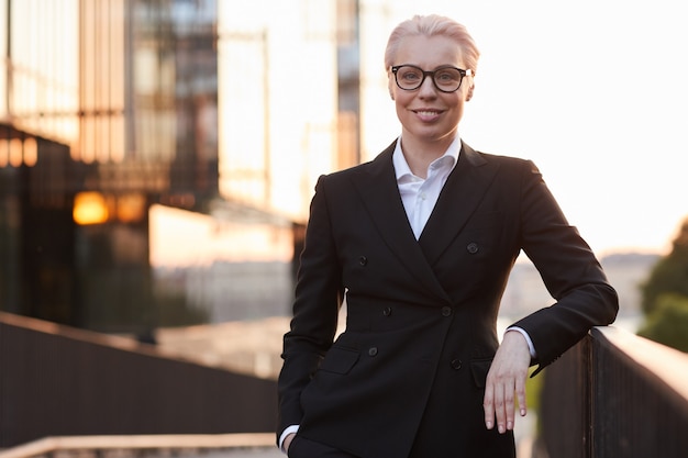 Portrait of mature businesswoman in eyeglasses and in black suit smiling at camera standing outdoors