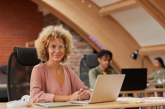 Photo portrait of mature businesswoman with curly hair in eyeglasses  while sitting at her workplace with laptop with her colleagues