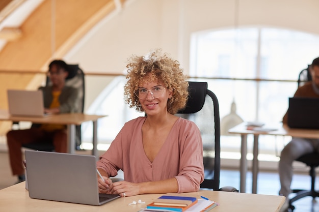 Portrait of mature businesswoman with curly hair sitting at her workplace with laptop and smiling at camera with her colleagues