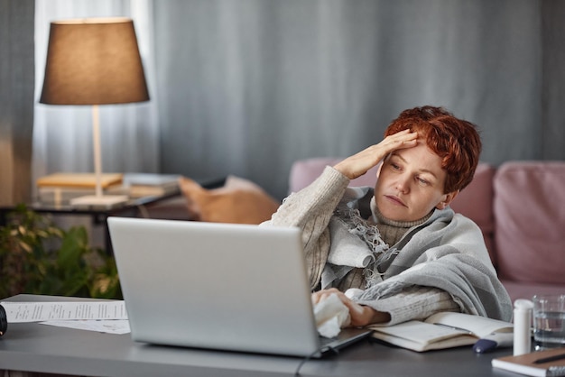 Portrait of mature caucasian woman having flu with severe headache wearing warm outfit sitting at de