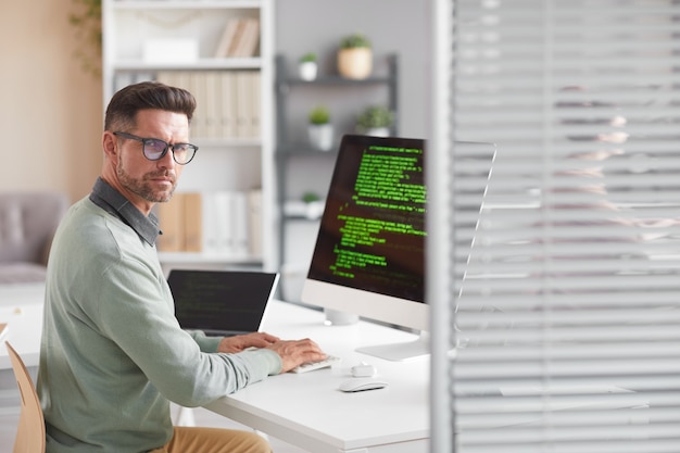 Portrait of mature developer looking at front while working at the table with computer in software developing company