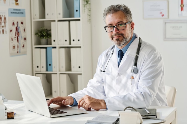 Portrait of mature doctor in white coat and eyeglasses looking at camera while working on laptop at