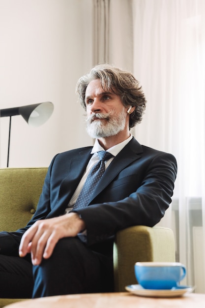 Portrait of a mature grey-haired businessman posing indoors at home near window dressed in formal clothes using earphones sitting on chair.