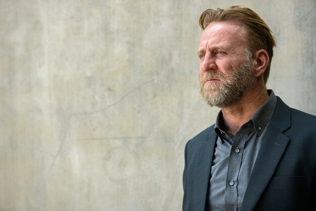 Portrait of mature handsome bearded businessman with blond hair in suit against concrete wall outdoors