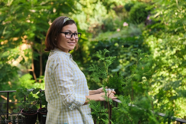 Portrait of mature housewife woman, female with cup of coffee on open balcony decorated with green plants, copy space