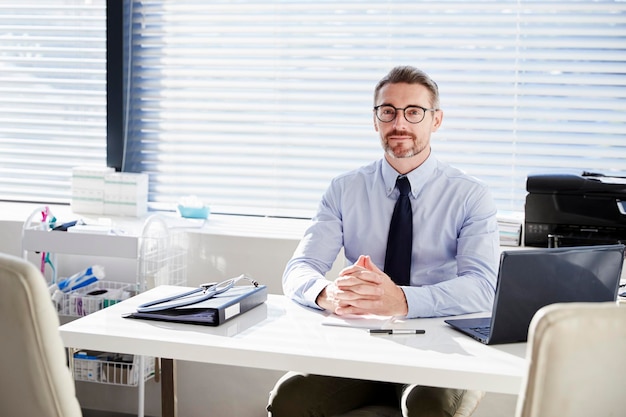 Portrait Of Mature Male Doctor Sitting Behind Desk In Office