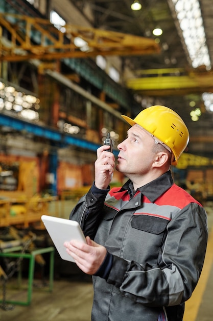 Portrait of mature manual worker in helmet and workwear looking at camera while using tablet pc in the factory