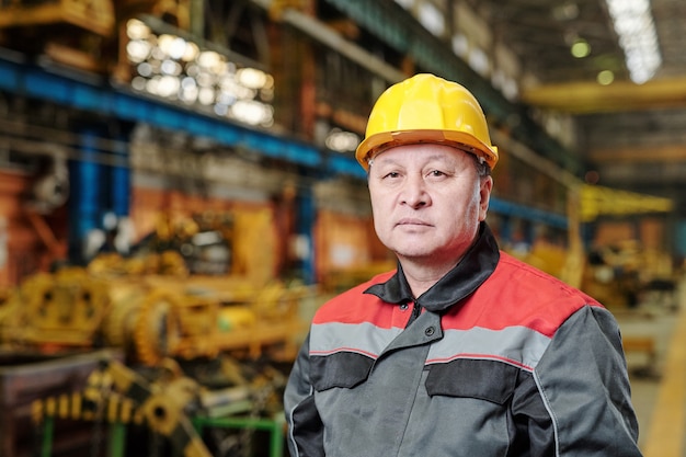 Портрет зрелого рабочего в шлеме и спецодежде, смотрящего в камеру во время работы на фабрике