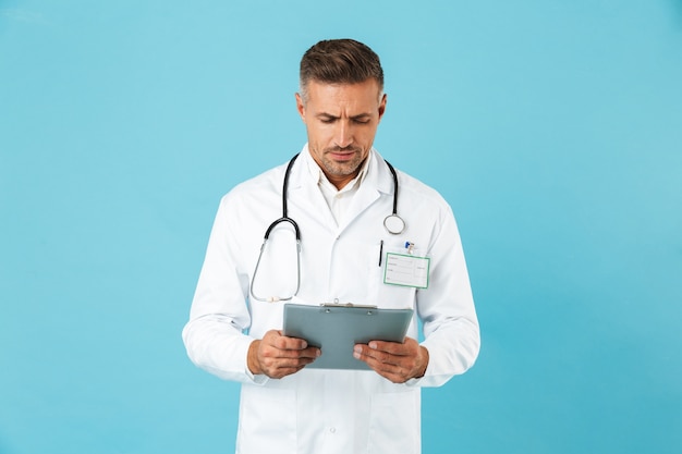 Portrait of mature medical doctor with stethoscope holding health card, standing isolated over blue wall