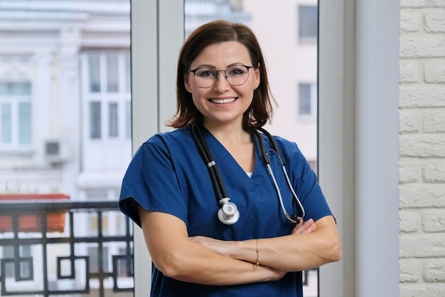 Portrait of mature nurse woman in blue uniform with stethoscope, smiling confident female looking at camera with arms crossed, medic in hospital near window